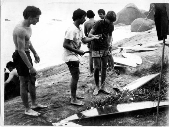 Durante o primeiro campeonato de surf na Trindade em 1977, Charlys Brown observa o Madureira entregando uma cordinha M&M, que ele fabricava a Roberto Moura, Trindade (RJ). Foto: Andy Goldstein.
